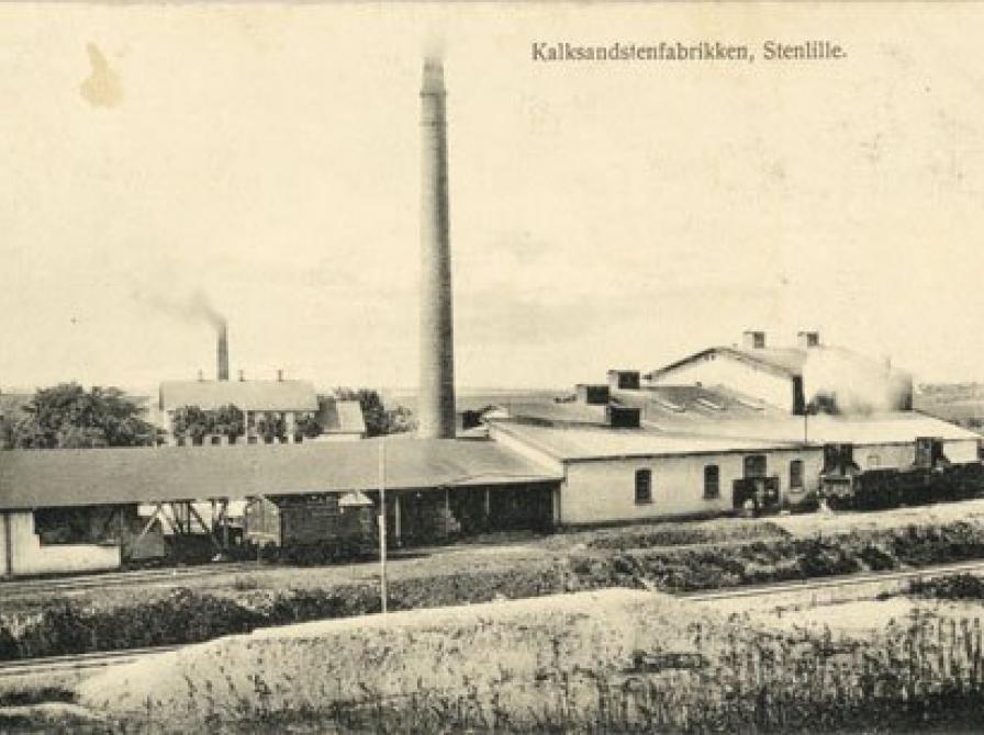 foto af Kalksandstensfabrikken med en stor skorsten. I forgrunden Høng-Tølløsebanens skinnelegeme fra 1901.